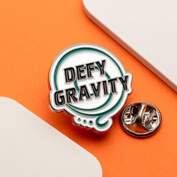 Defy Gravity lapel pin with black text and green swirling graphic on a white background.
