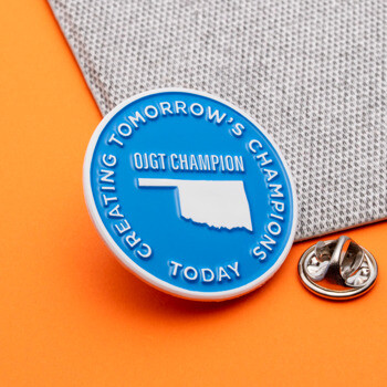 Circular blue lapel pin with white Oklahoma silhouette and OJGT Champion text on an orange and grey background.