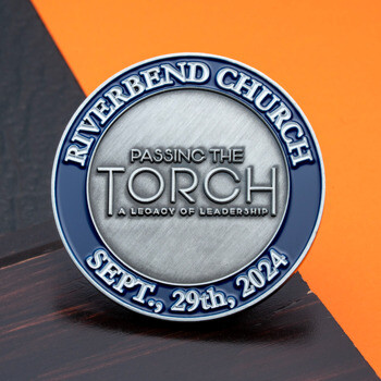 Circular challenge coin for Riverbend Church with blue enamel border and brushed silver center reading Passing the Torch Sept. 29th, 2024.