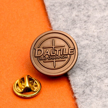 Round antique bronze Daltile R&D Champion lapel pin shown with a gold butterfly clutch backing on an orange and grey surface.